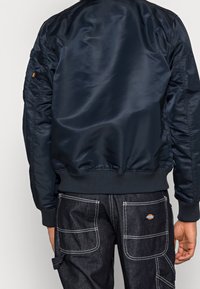 Giacca bombardiere navy con finitura in satin lucido, polsini e bordo a costine, e una tasca laterale. Indossata con pantaloni cargo in denim scuro con cuciture bianche.