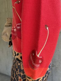 Piros kötött pulóver, melyen hímzett cseresznye motívumok találhatók az ujjakon. Leopárdmintás alsó szegéllyel és sima textúrával díszítve.