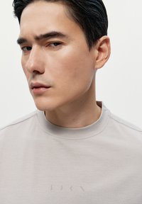 Šedé tričko s kulatým výstřihem a minimalistickým designem, s jemným brandingem. Hladký povrch s mírně melírovaným vzhledem.