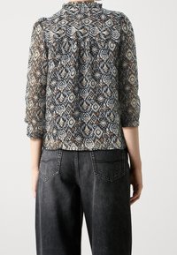 Blouse ample en tissu transparent avec un col haut, ornée d'un motif de losanges bleu, marron et crème. Associée à un jean sombre.