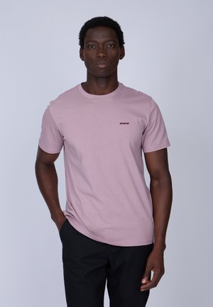T-shirt rose à manches courtes en tissu doux, avec un col rond et un détail brodé minimaliste. Le modèle porte un pantalon noir à rayures ajusté.