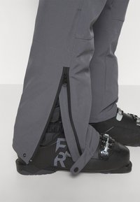 Szare spodnie narciarskie z bocznymi zamkami, elastycznymi mankietami i zwężanym krojem, w połączeniu z czarnymi butami narciarskimi z metalowymi klamrami i dobrą przyczepnością.