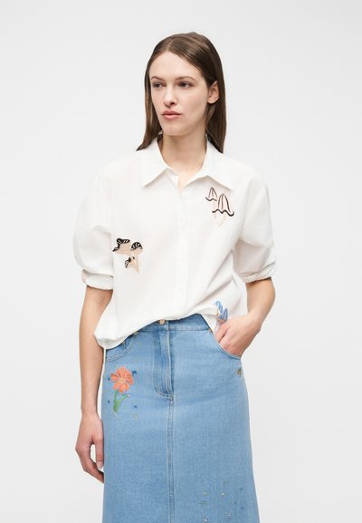 Kvinne iført en hvit skjorte med soppsømsøm og et blått denimskjørt med blomster- og stjernesøm, stående med en hånd i lommen.