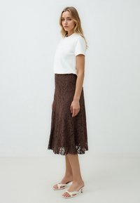 Brauner Spitzen-Midirock mit ausgestelltem Design, kombiniert mit einem weißen Kurzarmoberteil. Das Modell trägt weiße High-Heels-Sandalen.