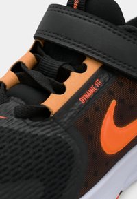 Μαύρο αθλητικό παπούτσι με υφή στην επάνω πλευρά, πορτοκαλί λεπτομέρειες και ιμάντα με Βέλκρο. Ξεχωρίζει για το αναπνεύσιμο πλέγμα και το δυναμικό σχεδιασμό εφαρμογής.