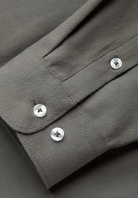 Manche de chemise grise à boutons, avec deux boutons ronds en argent et des coutures contrastantes. Le tissu semble doux avec une légère texture.