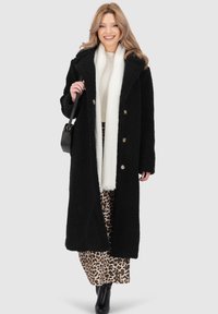 Donna sorridente, con un lungo cappotto nero texturizzato, sciarpa bianca, gonna con stampa leopardata, stivali neri e in mano una borsa nera su sfondo grigio.