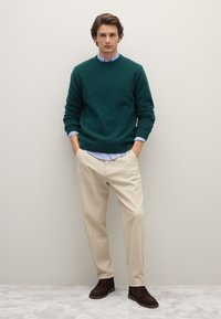 Dunkelgrüner, strukturierter Pullover über einem hellblau-weiß gestreiften Hemd, kombiniert mit beigen Hosen und dunkelbraunen Schuhen. Einfaches, figurbetontes Design.