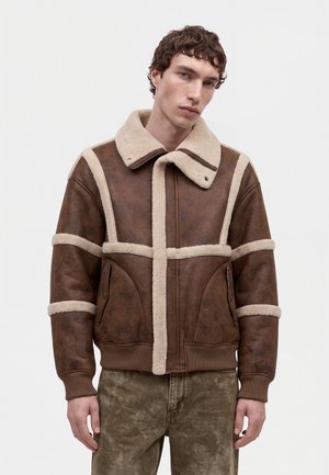 Un mannequin masculin porte une veste marron doublée de peau lainée et un pantalon olive sur un fond clair et uni.