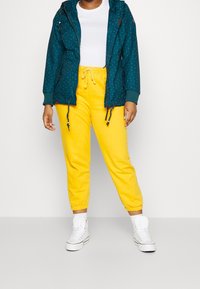 Teal jacket with white polka dots, drawstring hood, and zip pockets. Bright yellow joggers with elastic cuffs. White high-top sneakers.