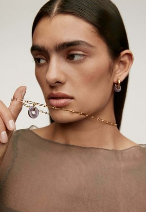 Collar de cadena de oro con eslabones, que presenta un colgante circular púrpura. El modelo lleva un aro a juego, ambos acentuando el tono de piel.