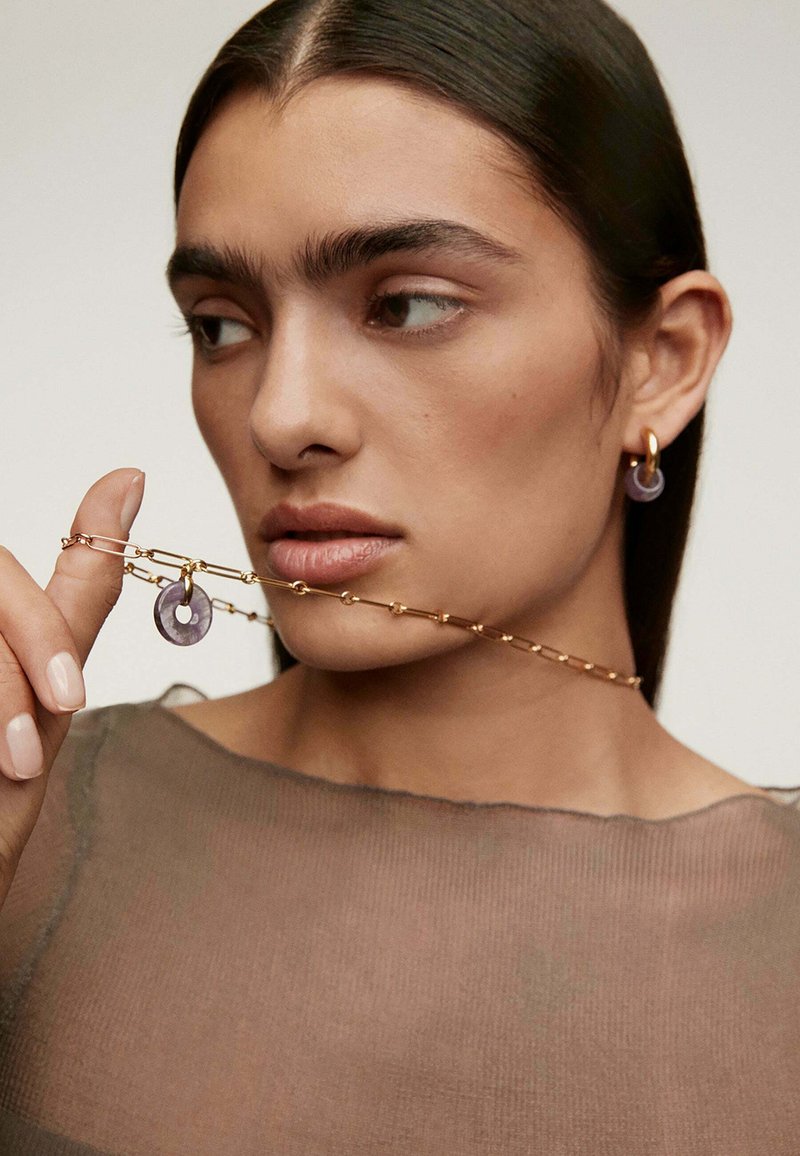 Collar de cadena de oro con eslabones, que presenta un colgante circular púrpura. El modelo lleva un aro a juego, ambos acentuando el tono de piel.