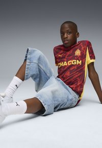 Röd och gul t-shirt med kort ärm och abstrakt mönster, tillsammans med ljusblå baggy denimshorts. Vita sneakers och strumpor med logotyp.