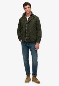 Veste olive avec plusieurs poches, assortie à une chemise à carreaux, un jean bleu et des bottes beiges. Le mannequin se tient de face.