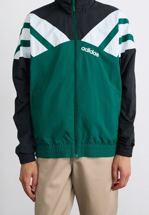 Veste zippée Adidas verte, noire et blanche avec poignets et ourlet élastiques portée avec un pantalon beige, montrant le haut du corps et les bras.