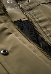 Gros plan sur une veste vert olive ouverte, montrant la fermeture éclair, la doublure en maille noire, le bouton-pression et une étiquette en tissu avec le logo de la marque et des icônes indiquant un tissu respirant.