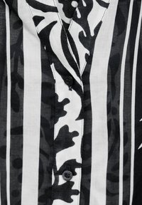 Camicia a maniche lunghe bianca e nera con strisce verticali e motivo camouflage. Il tessuto appare leggero e con una texture liscia.