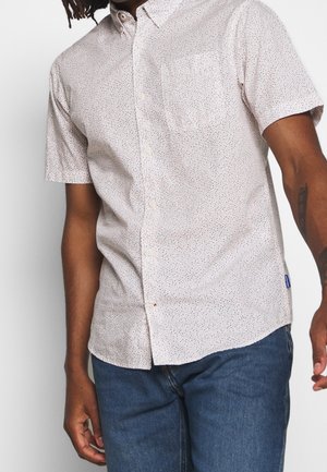 Mand iført en lysefarget, kortærmet skjorte med små prikker og blå jeans mod en ensfarvet baggrund.