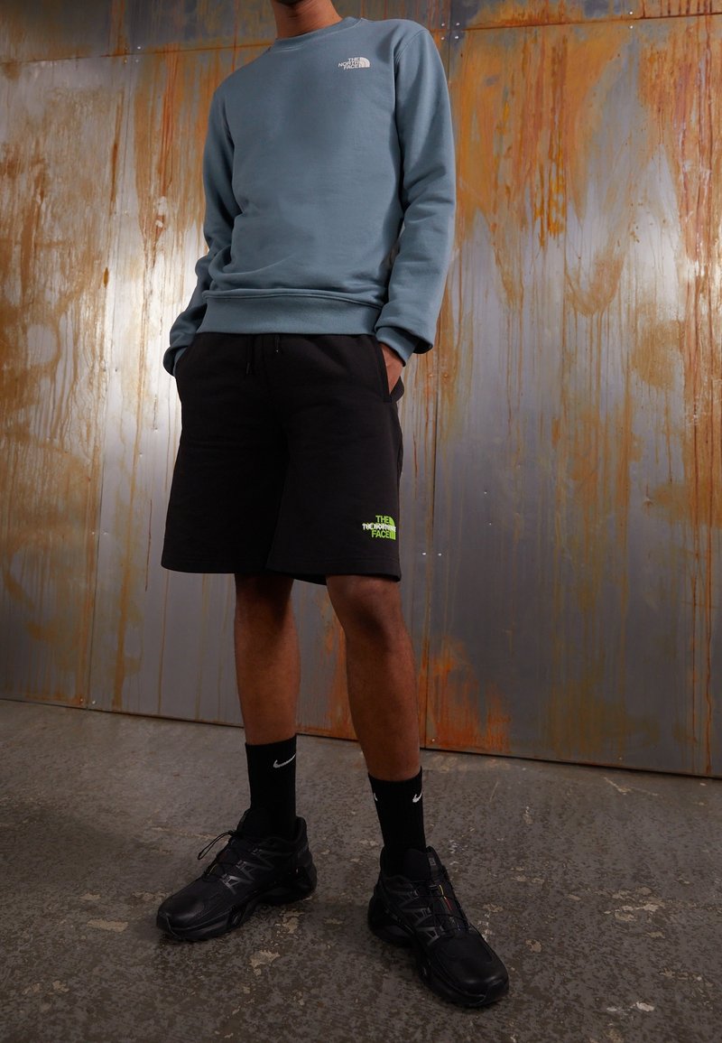 Světle modrá mikina, černé šortky s neonovým logem, černé tenisky a černé ponožky. Šortky mají elastický pas a kapsy.