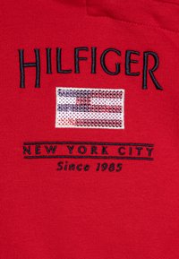 Tissu rouge avec broderie "HILFIGER" au-dessus d'un motif de drapeau pailleté et le texte "NEW YORK CITY", terminé par "Depuis 1985" en dessous.