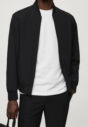 Giubbotto bomber leggero nero con zip, polsini a coste e colletto rialzato, indossato sopra una maglietta bianca. Tessuto testurizzato con design minimale.