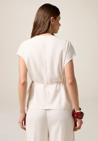 Blusa crema a maniche corte con vita arricciata, colletto tondo, texture liscia, abbinata a pantaloni larghi e braccialetti multicolori.