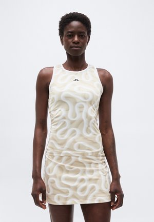 Ženska, oblečena v brezrokavno bež obleko z belim abstraktnim vzorcem v obliki vijug, stoji pred enobarvnim svetlim ozadjem.