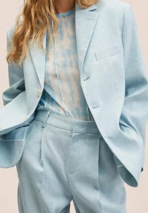 Mujer con traje sastre azul claro con blazer y pantalones plisados, combinado con una blusa transparente de patrón azul y blanco.