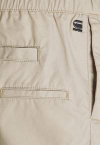 Beige fabric with two horizontal slit pockets and a small black "G RAW" logo near the elastic waistband.