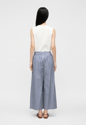 Žena s dlhými čiernymi vlasmi, oblečená v bielom tielku bez rukávov, modro-bielych zvislých pruhovaných širokých nohaviciach a hnedých sandáloch, stojaca chrbtom.