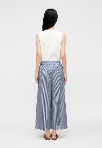 Femme aux longs cheveux noirs portant un haut blanc sans manches, un pantalon large à rayures verticales bleues et blanches, et des sandales marron, debout de dos.