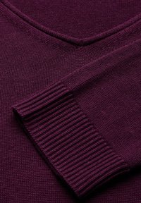 Plommonfärgad stickad tröja med V-ringning, där ena ärmen är vikt över bröstet och visar ribbade manschettdetaljer.