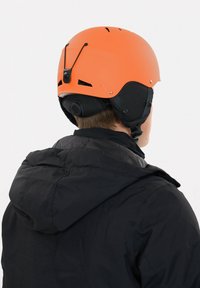 Orange sports hjelm med ventilationshuller, mat finish og justerbar hagerem, placeret mod en sort hættetrøje.