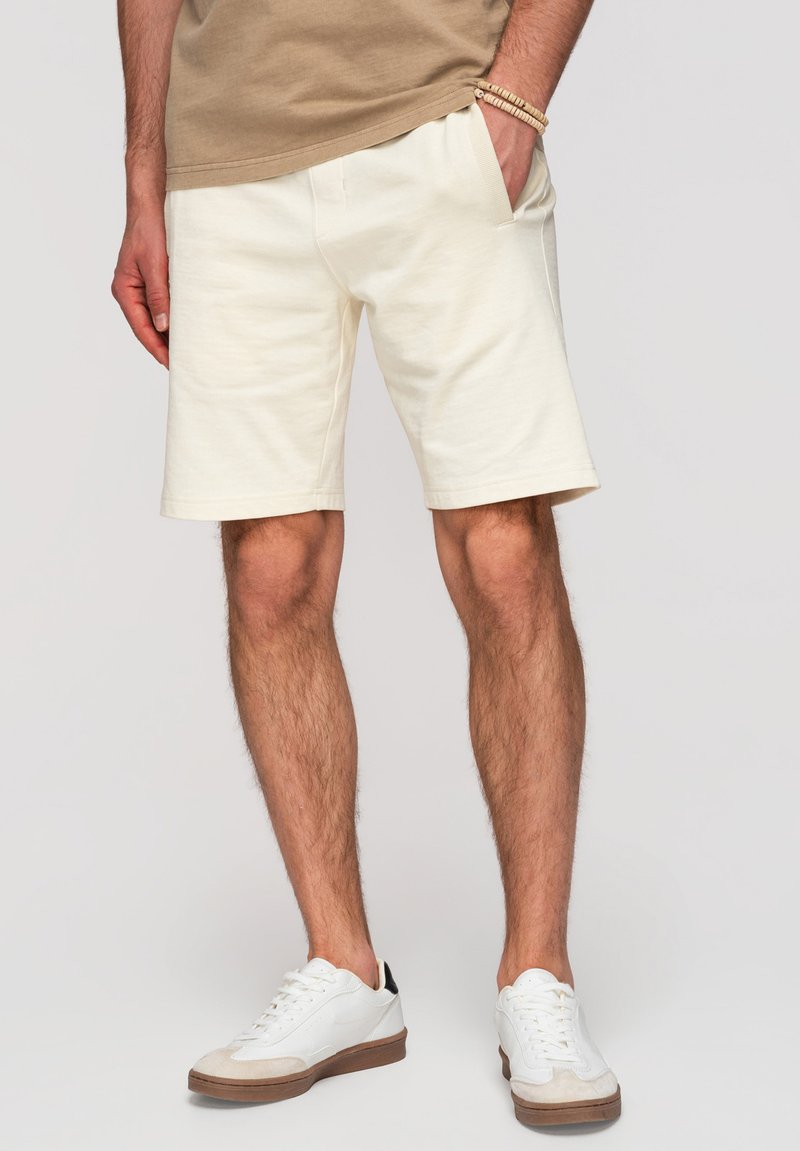 Homme portant un short beige, un t-shirt couleur sable, des baskets blanches et un bracelet en perles, debout avec une main dans la poche devant un fond uni.