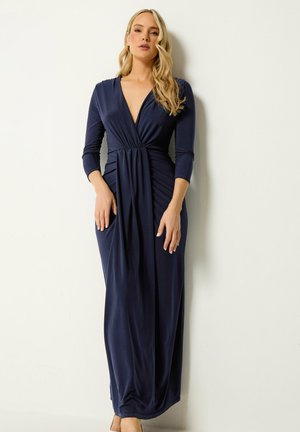 Frau mit langen blonden Haaren, die ein marineblaues, langärmliges Kleid mit tiefem V-Ausschnitt und bodenlanger Länge trägt, steht vor einer schlichten hellen Wand.