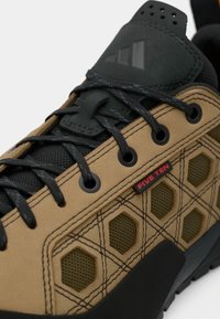 Καφέ και μαύρα παπούτσια πεζοπορίας με ανάγλυφη επιφάνεια, πάνελ από δίχτυ, εξαγωνικές οπές και μαύρα κορδόνια· διαθέτουν ανάγλυφο λογότυπο στη γλώσσα.