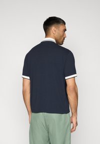 Μπλε ναυτικό πόλο μπλουζάκι με λευκές λεπτομέρειες στον γιακά και τις μανσέτες, με κοντά μανίκια και άνετη γραμμή, εμφανιζόμενο από πίσω. Ομαλή υφή βαμβακιού.