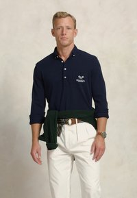 Navy long-sleeve polo with a collar and pocket detail. Green sweater tied at the waist, white trousers, brown belt, and watch accentuate the look.