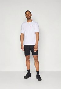 Camiseta blanca de algodón con un logo rojo en el pecho izquierdo, combinada con pantalones cortos negros y zapatillas negras, posando sobre un fondo neutro.