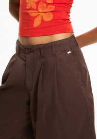Brown, loose-fitting, pleated trousers with a button closure and side pockets. Cotton material with a smooth texture.