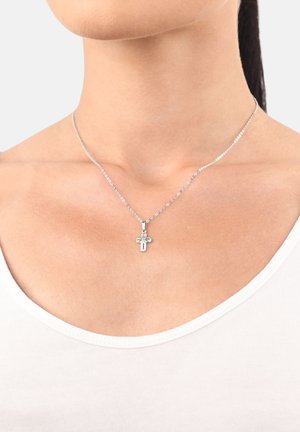 Collar de plata con una cadena delgada, que presenta un pequeño colgante en forma de cruz adornado con detalles sutiles, llevado sobre una blusa blanca.