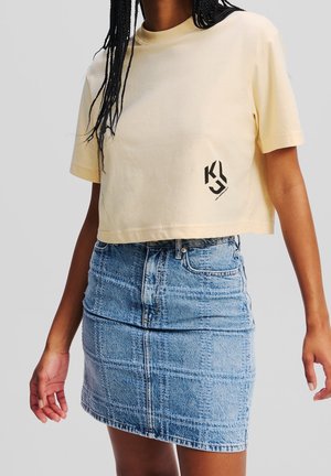 Femme portant un t-shirt court jaune pâle avec logo et une jupe en jean bleue taille haute avec poches avant et détails de couture.