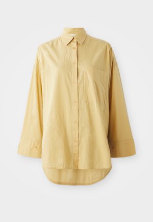 Chemise jaune à boutons, avec une poche, manches longues et ourlet arrondi. Fabriquée en tissu léger et texturé. Dotée de boutons oranges.