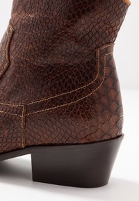 Bota de vaquero de cuero marrón con patrón de piel de serpiente en relieve y costura beige, con un tacón oscuro apilado sobre una superficie blanca.