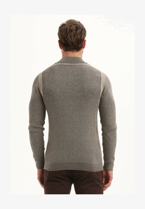 Vīrietis ģērbts pieguļošā ribotā pelēkā džemperī ar gaišākām pelēkām svītrām uz pleciem, skatīts no mugurpuses uz vienkārša fona.
