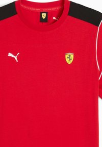 Cămașă sport roșie cu un panou negru pe umăr, logo Puma și un emblemă galbenă Ferrari cu un design al unui cal învățat.