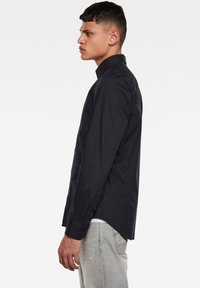 Camicia nera a maniche lunghe con colletto classico, design aderente e tessuto liscio. Presenta polsini con bottone e orlo curvo.