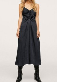 Musta maksimekko, jossa on V-niska ja rypytys rinnassa, virtaava siluetti, valmistettu sileästä kankaasta. Yhdistetty polveen ulottuviin mustiin saappaisiin.