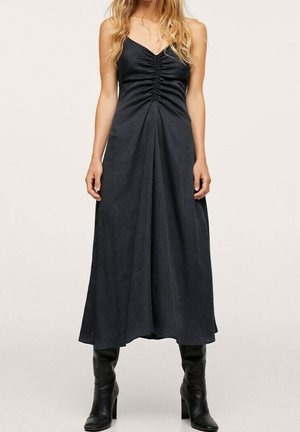 Zwarte maxi-jurk met een V-hals en plooien bij de buste, vloeiende silhouet, gemaakt van een gladde stof. Gemaakt in combinatie met zwarte laarzen tot de knie.