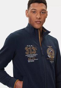 Giacca zip-up della marina realizzata in morbido materiale, con ricamo in oro e bianco, colletto alto e dettagli a contrasto in beige ai polsini.